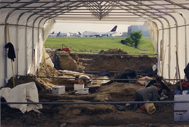 Archaeological Site People work at Elmbank Cemetery archaeological site under a canopy. A plane is in the background.